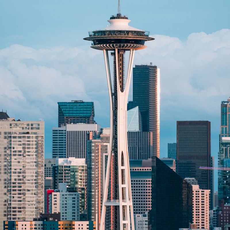 The image shows the Space Needle, a famous landmark, surrounded by several buildings in a city skyline under a partly cloudy sky.