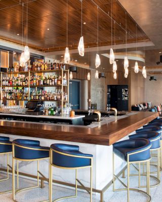 A modern bar with blue stools, a wooden countertop, and a fully stocked backlit shelf surrounded by a cozy seating area and unique lighting fixtures.