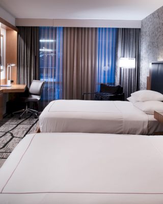 A well-lit hotel room with two neatly made beds, a flat-screen TV on the wall, a desk with a chair, modern decor, and large windows with curtains.