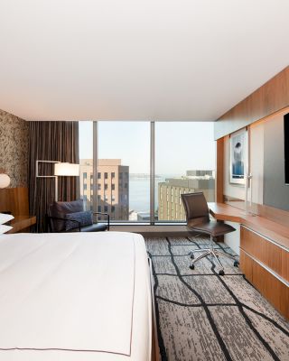 A modern hotel room with a large bed, wall-mounted TV, desk, chair, and floor-to-ceiling windows offering a cityscape view and plenty of natural light.