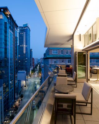 A modern balcony overlooking a cityscape with tall buildings, featuring outdoor seating and tables, under a partially covered area in the evening.
