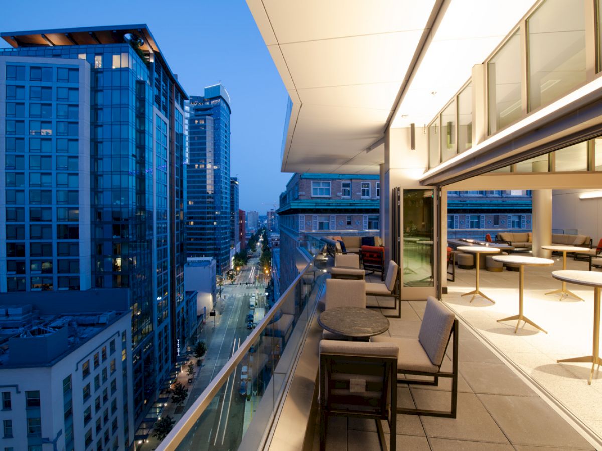 The image shows a modern balcony with tables and chairs, overlooking a city street lined with high-rise buildings at dusk.