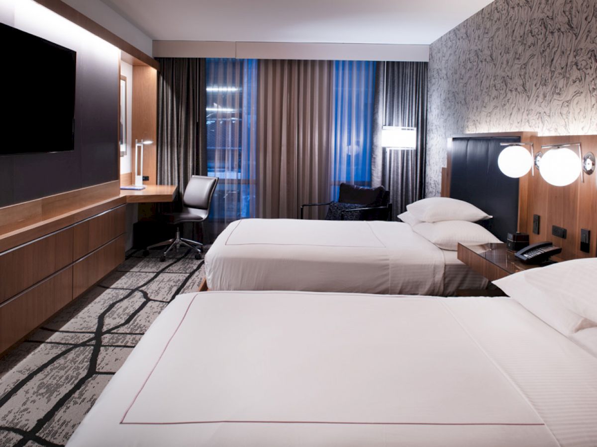 A modern hotel room with two double beds, a TV on the wall, a desk with a chair, bedside lamps, and large windows with curtains.
