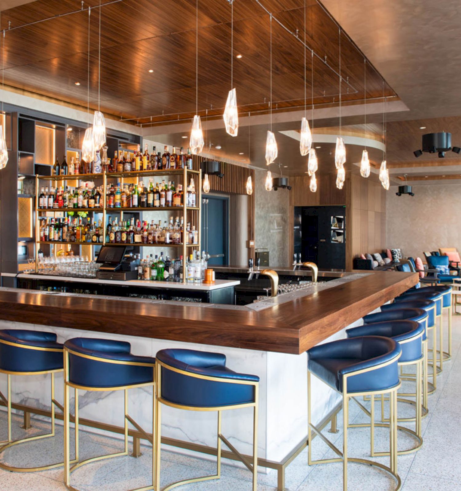 The image shows a modern bar with blue chairs, a wooden countertop, and shelves stocked with various bottles, located in a spacious, chic interior.