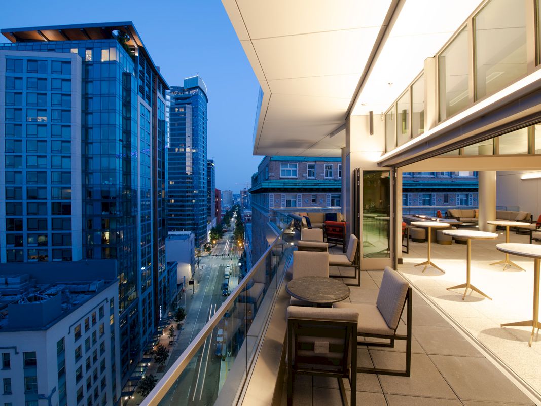 A modern rooftop terrace overlooking a city street, with seating areas and high-rise buildings illuminated at dusk.