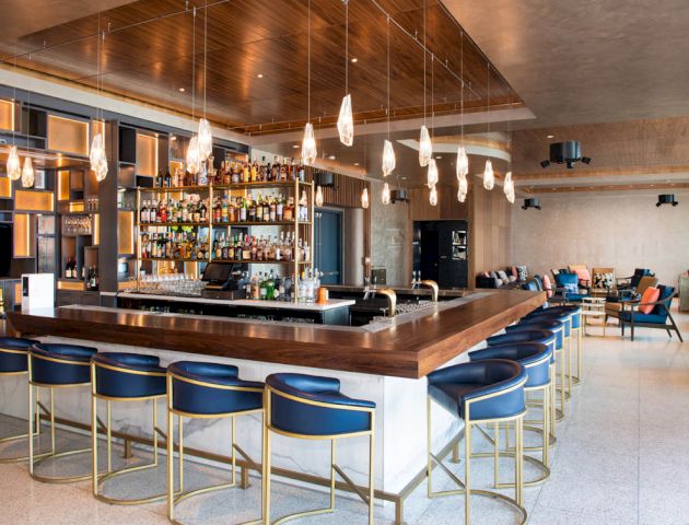 This image shows a modern bar with a long counter, blue stools, a large selection of bottles on shelves, and pendant lights hanging from the ceiling.