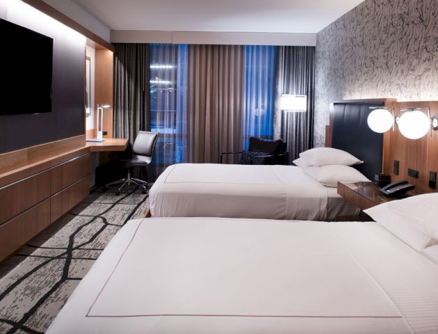 A modern hotel room with two queen beds, wall-mounted TV, desk with a chair, floor lamps, and curtains on the windows, all in a contemporary design.