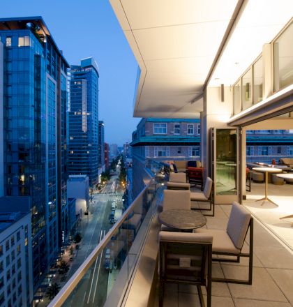 A modern rooftop terrace overlooking city skyscrapers and a street below, featuring outdoor seating and tables.