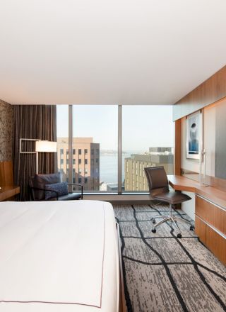 A modern hotel room with a large bed, a wall-mounted TV, a desk, and floor-to-ceiling windows offering a city and water view.