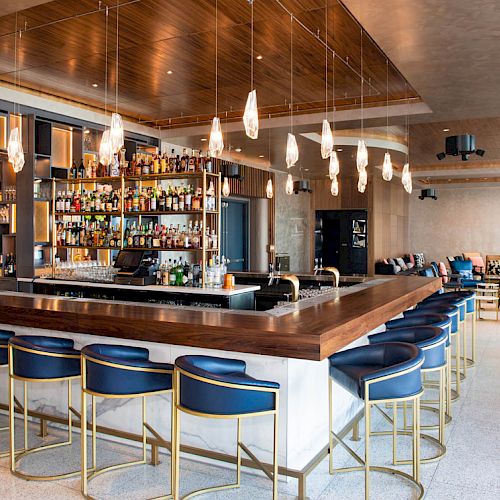 A modern bar features stylish lighting, a well-stocked shelf, blue stools with gold accents, and a spacious seating area with contemporary decor.