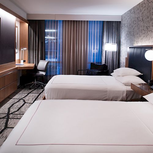 The image shows a modern hotel room with two double beds, a flat-screen TV, a work desk, and contemporary decor.