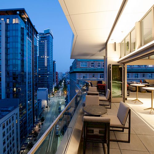 The image shows a modern cityscape at dusk with high-rise buildings and a well-lit rooftop terrace featuring outdoor seating and tables.