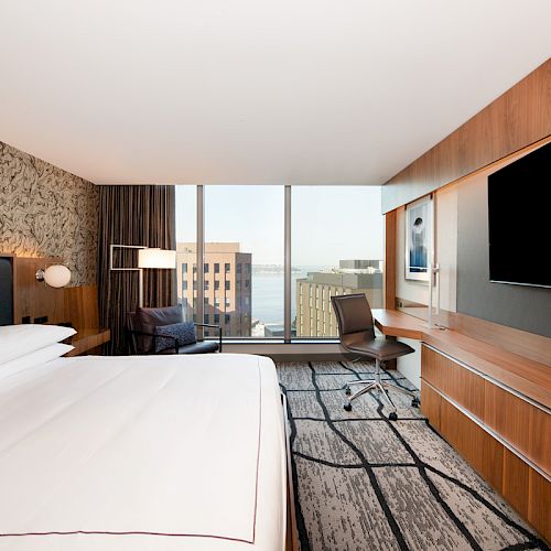 A modern hotel room features a large bed, wall-mounted TV, desk with chair, and a window offering a cityscape view with buildings and water.