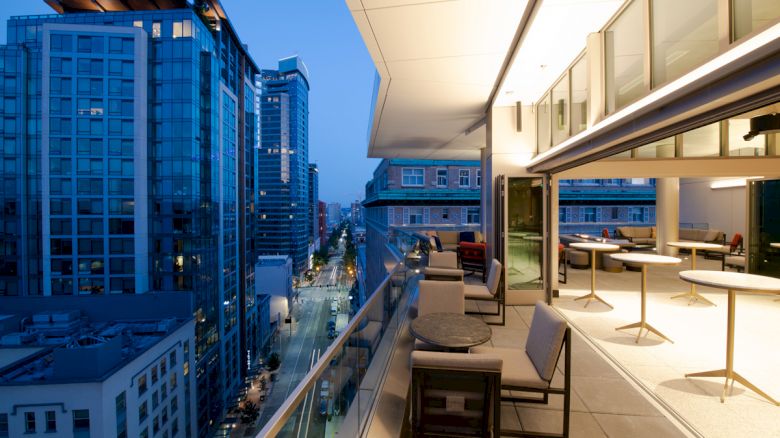 A luxurious rooftop patio with modern furniture overlooks a cityscape at dusk, featuring towering buildings and a lit street below.