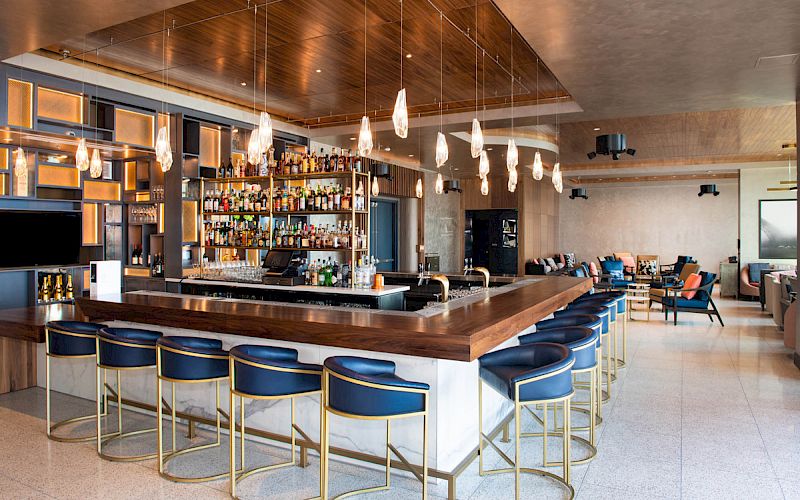 The image shows a modern bar with a wooden counter, blue and gold bar stools, a well-stocked liquor shelf, and contemporary lighting fixtures.