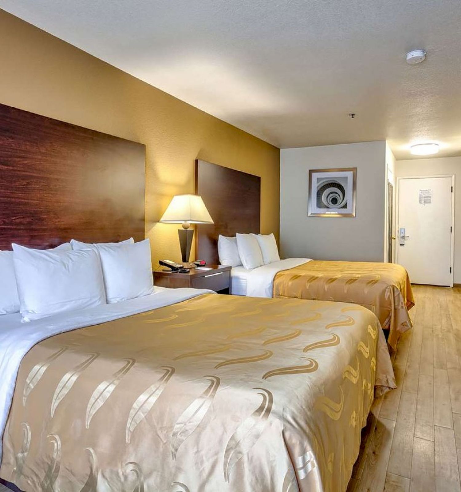 The image shows a hotel room with two double beds, bedside tables, a lamp, a TV on a dresser, and a kitchenette area. The room has wooden flooring.