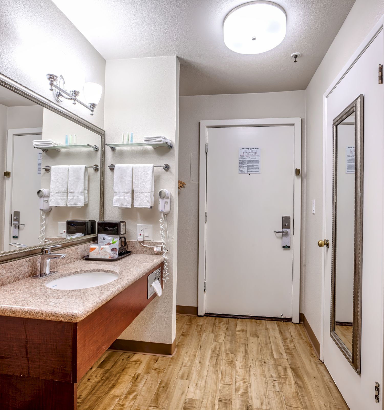 The image shows a hotel bathroom with a sink, large mirror, towels, and a door with a full-length mirror. The floor is wood, and the lighting is bright.