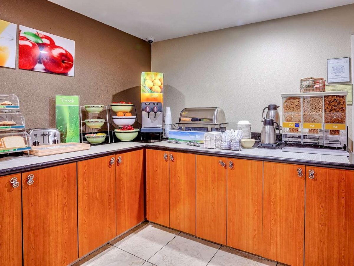 This image shows a breakfast buffet setup with cereals, bread, fruit, juice, and coffee on a wooden counter in a brightly lit room.