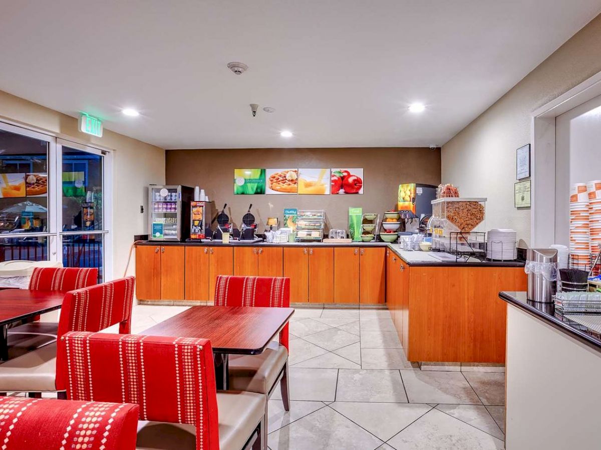 The image shows a small dining area with red chairs and tables, a breakfast bar with various food and drink items, and bright lighting.