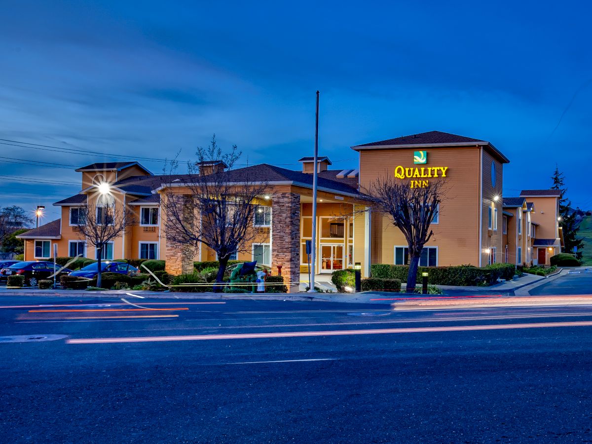 An illuminated building with a "Quality Inn" sign is pictured at dusk, with light trails from passing cars on the street in front of it.