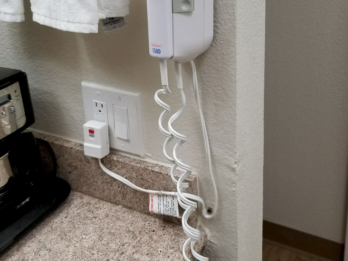 A wall-mounted hairdryer with a coiled cord is beside a countertop with a coffee maker, outlet, and power switch in what appears to be a hotel room.