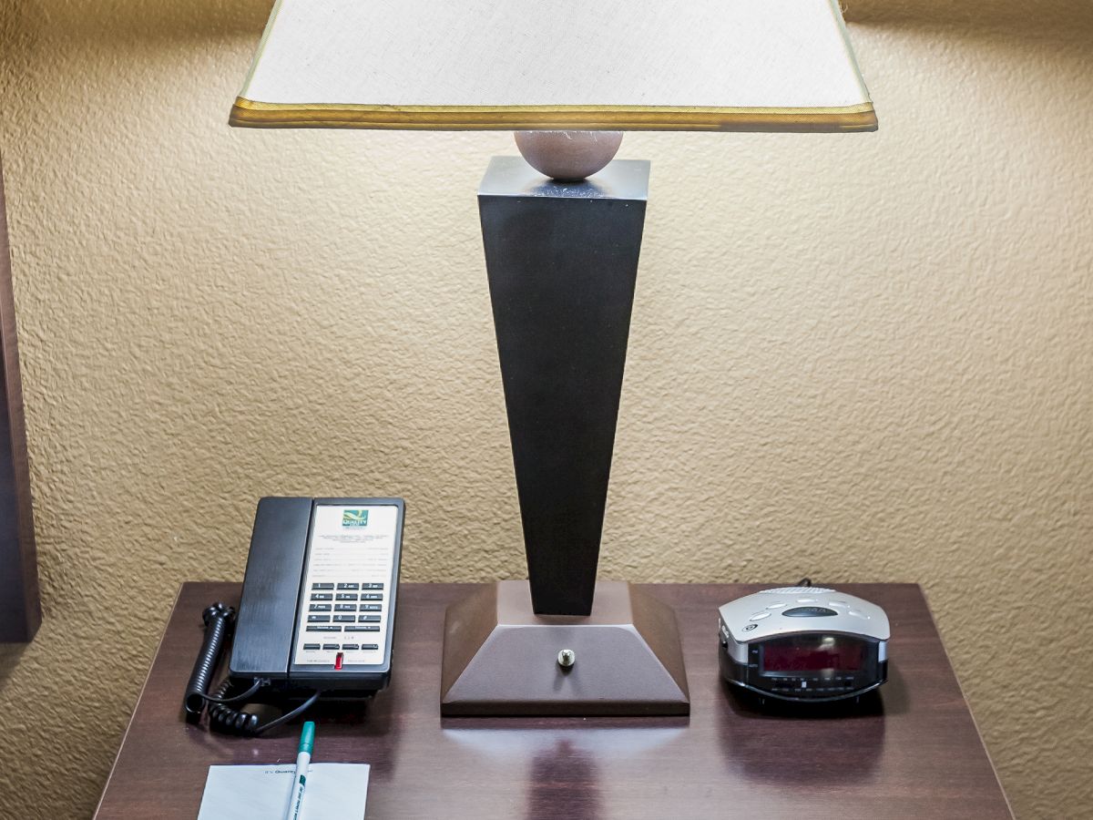 A bedside table with a lamp, a telephone, a notepad with a pen, and a digital clock are arranged on it.