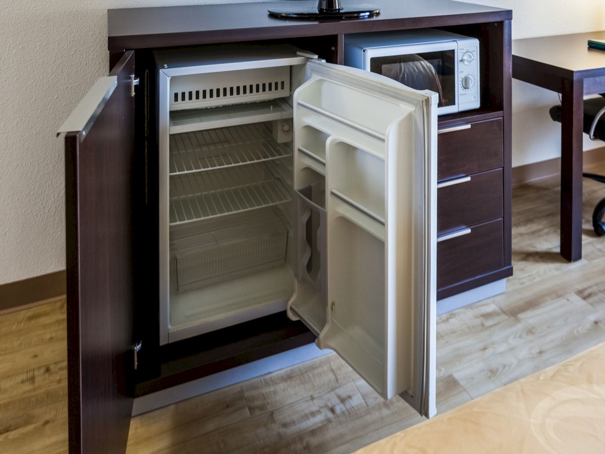 An open mini-fridge embedded within a wooden cabinet, with a microwave on top and a desk and chair visible in the background.