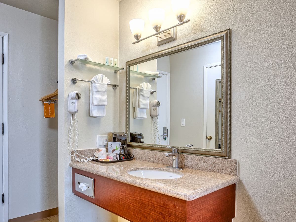 The image shows a well-lit bathroom vanity area with a sink, mirror, towels, a hairdryer, and various toiletries on the countertop.