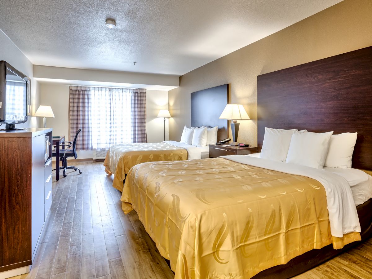 The image shows a hotel room with two double beds, a TV, a desk, and a window with curtains in the background.