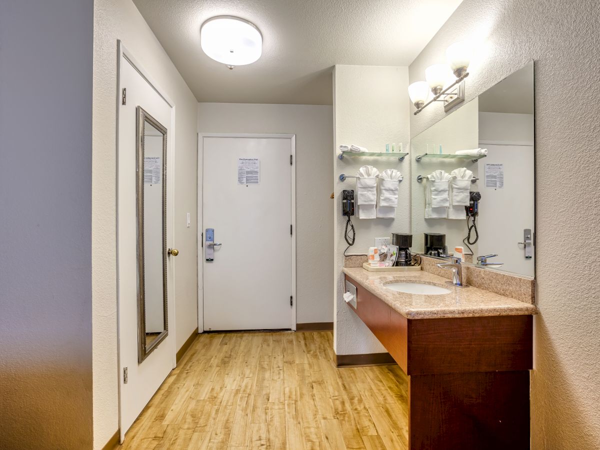 A hotel room entryway with a vanity area, large mirror, sink, coffee maker, hanging towels, and wooden floor, leading to a white door.